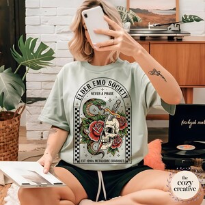 May include: A light green t-shirt with a graphic featuring a skull, snake, roses, and the text "ELDER EMO SOCIETY NEVER A PHASE". The design includes the text "EST 1990s METALCORE CRASHOUT". The shirt is a casual wear item.