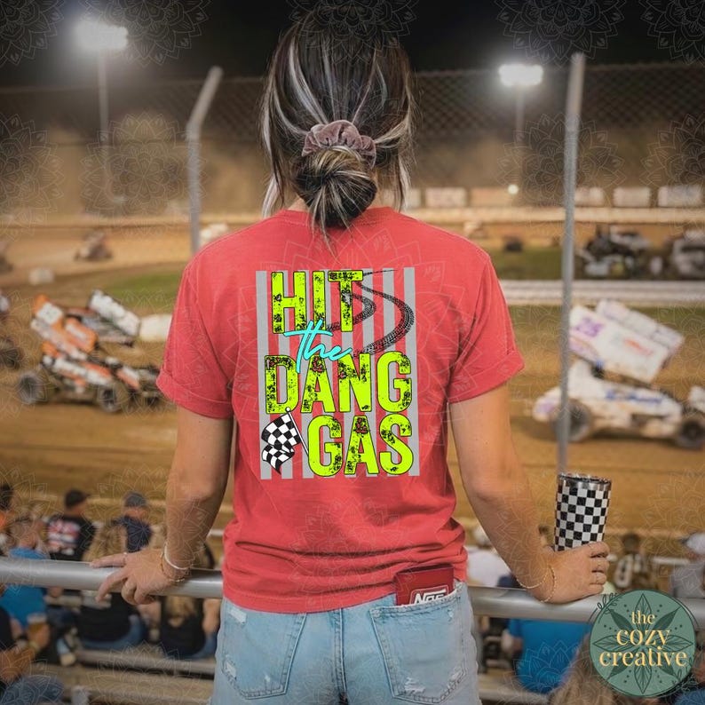 May include: Red t-shirt with the text "Hit the Dang Gas" in neon green and blue. The shirt features a checkered flag graphic and a tire track design. The person is holding a checkered tumbler.