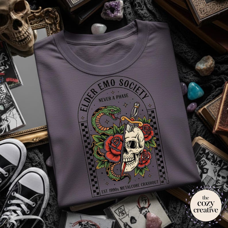 May include: A purple crew neck t-shirt featuring a graphic design with a skull, snake, roses, and a dagger. The text reads "ELDER EMO SOCIETY NEVER A PHASE" and "EST 1990'S METALCORE CRASHOUT".