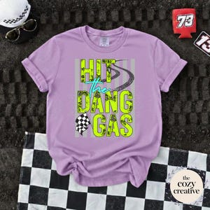 May include: A lilac t-shirt featuring the neon green and teal text "HIT the DANG GAS", with tire tracks and a checkered flag design. The image also shows sunglasses, a hat, and a checkered flag.