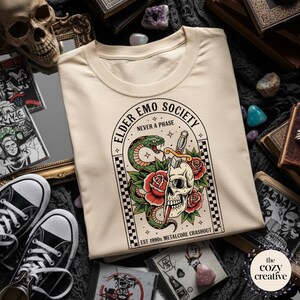 May include: Cream-colored t-shirt featuring a graphic with a skull, snake, roses, and the text "ELDER EMO SOCIETY NEVER A PHASE". The design includes a checkerboard border and the text "EST 1990s METALCORE CRASHOUT".