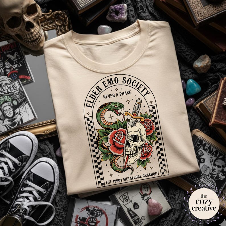 May include: Cream-colored t-shirt featuring a graphic with a skull, snake, roses, and the text "ELDER EMO SOCIETY NEVER A PHASE". The design includes a checkerboard border and the text "EST 1990s METALCORE CRASHOUT".