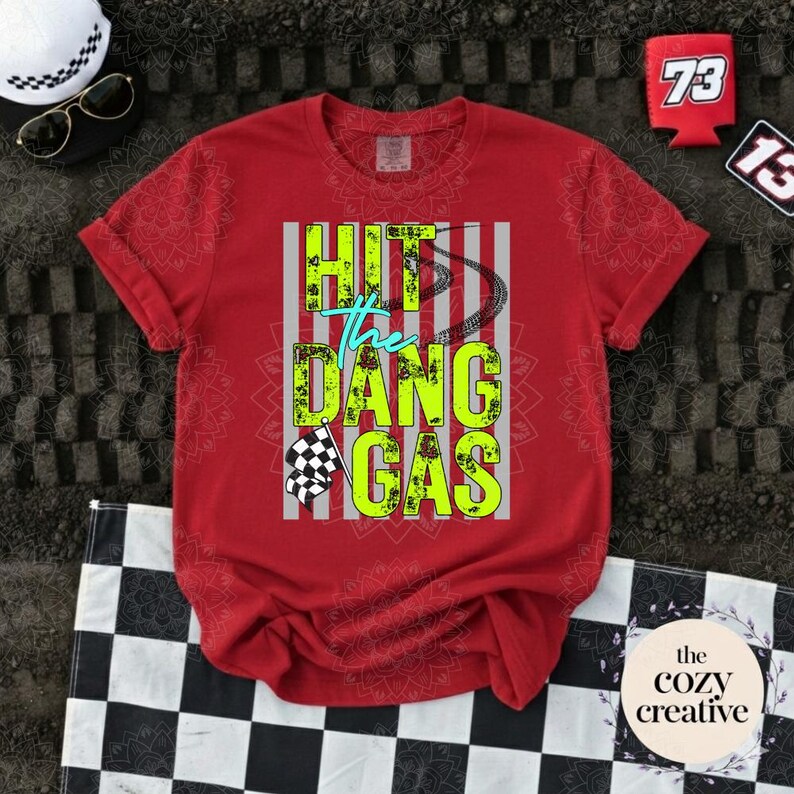 May include: Red t-shirt featuring the text "HIT the DANG GAS" in neon green and blue. The design includes a checkered flag and a motorcycle chain. The shirt has short sleeves and a crew neck.