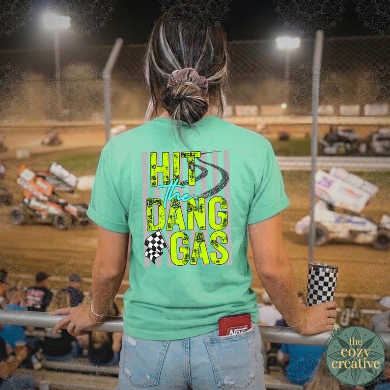 May include: A light green t-shirt with the text "Hit the Dang Gas" in a neon green and blue design. The shirt features a checkered flag graphic and a race track design. The person is wearing blue jeans and holding a checkered tumbler.