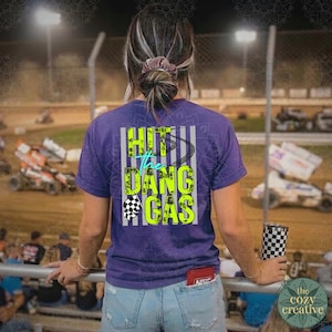 May include: A purple t-shirt featuring the text "Hit the Dang Gas" in neon green and teal, with a checkered flag and tyre track graphic. The shirt is worn by a person at a race track.