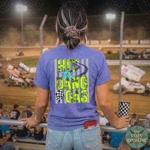 May include: A lilac t-shirt featuring the text "Hit the Dang Gas" in neon green and teal, with a checkered flag and tyre tracks. The shirt is worn by a person at a race track.