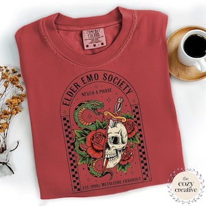 May include: A red Comfort Colors t-shirt with a graphic featuring a skull, snake, roses, and the text "ELDER EMO SOCIETY NEVER A PHASE". The design includes a sword and the text "EST 1990s METALCORE CRASHOUT".