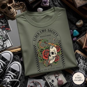 May include: An olive green t-shirt featuring a graphic design with a skull, snake, roses, and a dagger. The text "ELDER EMO SOCIETY NEVER A PHASE" and "EST 1990s METALCORE CRASHOUT" are also visible.