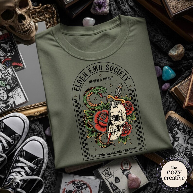 May include: An olive green t-shirt featuring a graphic design with a skull, snake, roses, and a dagger. The text "ELDER EMO SOCIETY NEVER A PHASE" and "EST 1990s METALCORE CRASHOUT" are also visible.