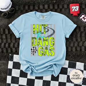 May include: A pale blue t-shirt featuring the text "HIT the DANG GAS" in neon green and teal. The design includes tyre tracks and a chequered flag. The shirt is folded on a chequered flag.