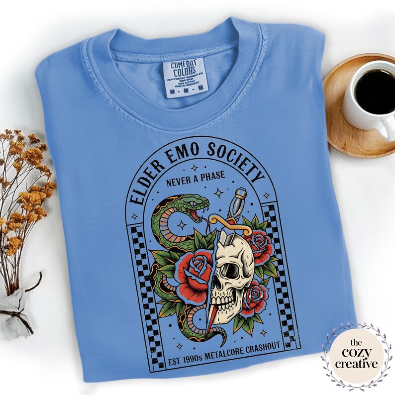 May include: A light blue Comfort Colors t-shirt featuring a graphic with the text "ELDER EMO SOCIETY NEVER A PHASE". The graphic includes a snake, skull, roses, and a dagger. The text "EST 1990s METALCORE CRASHOUT" is also present.
