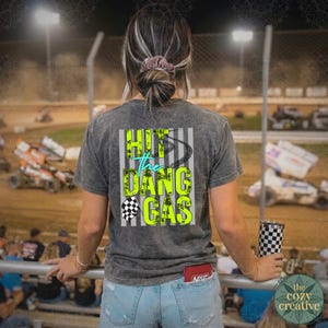 May include: A grey t-shirt with the text "Hit the Dang Gas" in neon green and teal, featuring a checkered flag and tyre track design. The shirt is worn by a person at a race track, holding a checkered tumbler.