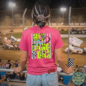 May include: A pink t-shirt with the text "Hit the Dang Gas" in neon green and teal, with a checkered flag graphic. The shirt is worn by a person at a race track, holding a checkered tumbler.