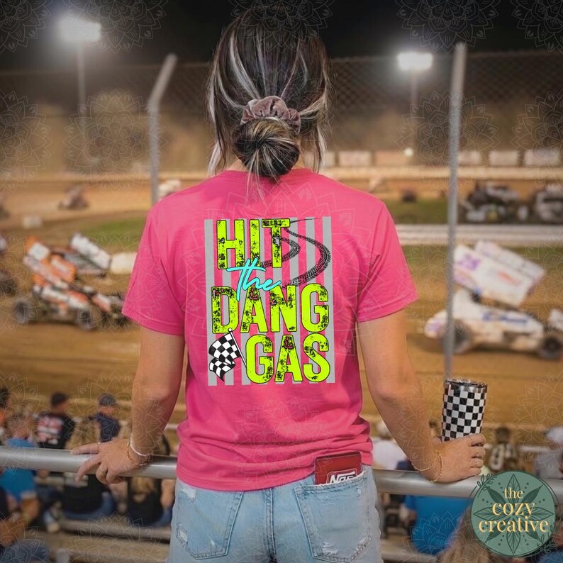 May include: A pink t-shirt with the text "Hit the Dang Gas" in neon green and teal, with a checkered flag graphic. The shirt is worn by a person at a race track, holding a checkered tumbler.