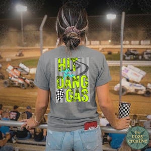 May include: A grey t-shirt with the text "Hit the Dang Gas" in neon green and teal, and a checkered flag graphic. The shirt is worn with light blue denim shorts. A checkered tumbler is held in the hand.