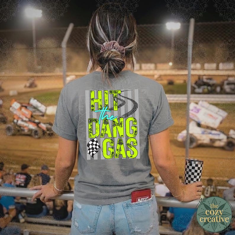 May include: A grey t-shirt with the text "Hit the Dang Gas" in neon green and teal, and a checkered flag graphic. The shirt is worn with light blue denim shorts. A checkered tumbler is held in the hand.