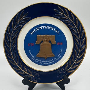 Può includere: Un piatto decorativo con bordo blu scuro e dettagli dorati. Il centro presenta la scritta "BICENTENNIAL", la Campana della Libertà e gli anni 1776 e 1976. Il piatto presenta anche l'iscrizione "PROCLAMATE LA LIBERTÀ IN TUTTA LA TERRA A TUTTI GLI ABITANTI".