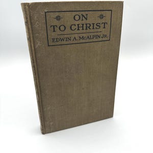 Peut inclure: Un livre vintage intitulé "ON TO CHRIST" par Edwin A. McAlpin Jr. Le livre a une couverture beige avec des lettres noires et un design rectangulaire. Le livre est debout sur un fond blanc.