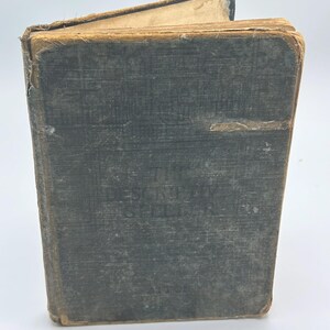 May include: An antique, weathered book with a dark gray, textured fabric cover. The front cover features the embossed title The Descriptive Speller. The edges of the book are frayed and worn, showing tan-colored binding and aged, yellowed paper pages.