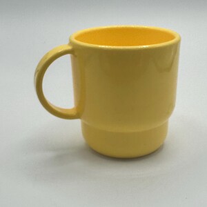 May include: A bright yellow ceramic mug with a smooth, glossy finish. The mug features a rounded, cylindrical body with a distinct horizontal ridge near the base and a curved handle attached to the side, providing a simple and functional design.