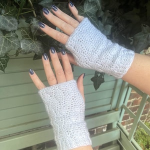 Könnte beinhalten: Ein Paar hellgraue, fingerlose Handschuhe. Die Handschuhe sind aus einem Strickmaterial mit einem strukturierten Muster gefertigt. Der Hintergrund zeigt grünes Laub und eine hellgrüne Holzoberfläche.