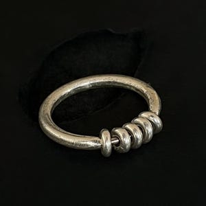 May include: A silver ring with a hammered texture and a series of small, connected rings. The ring is set against a dark background, highlighting its metallic sheen and simple design.