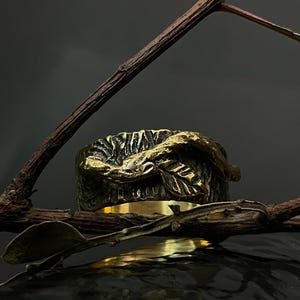 May include: A bronze-colored ring with a detailed leaf design. The ring is set against a dark background, with a branch and leaves in the foreground. The ring's band is a polished gold color.