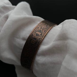 May include: A copper cuff bracelet with a geometric design. The bracelet, with its repeating shapes and lines, is wrapped around white fabric. The pattern creates a textured appearance. The bracelet's colour is a warm copper.