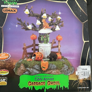 May include: A Halloween-themed table accent featuring a tree with hanging ghost lights, a cat, and pumpkins. A green ghoul emerges from a trash can. The base includes a fence, a bear, and a jack-o'-lantern. The text reads "GARBAGE GHOUL".