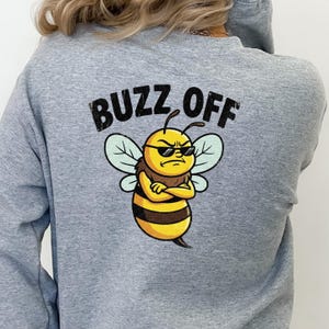 May include: A light gray sweatshirt with the words "BUZZ OFF" printed across the top. Below the text is a cartoon bee wearing sunglasses with its arms crossed. The bee is yellow and brown with green wings.