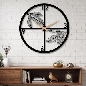 Puede incluir: Un reloj de pared circular con un marco de metal negro y un diseño recortado de ramas con hojas. La esfera muestra los números 3, 6, 9 y 12 en una fuente negra en negrita. Dos manecillas de madera clara indican la hora sobre la estructura.
