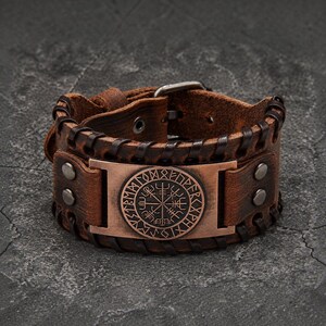 May include: A wide brown leather cuff bracelet featuring a central copper-toned rectangular plate with a circular Vegvisir symbol surrounded by runic characters. The leather band is secured with dark brown lacing and metal rivets.
