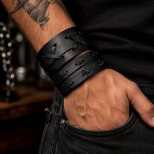 May include: A wide black leather cuff bracelet featuring a series of decorative crisscross lacing details along the center. The bracelet is constructed from multiple layered bands of black leather, secured with matching black stitching and lacing.