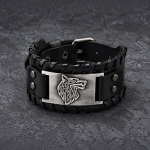 May include: A black leather cuff bracelet featuring a rectangular silver-toned metal plate with an embossed wolf head design. The leather band is detailed with stitched edges and secured by two metal rivets on each side and a metal buckle closure.