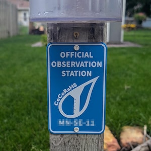 May include: A blue sign reading "OFFICIAL OBSERVATION STATION" with a white logo and text, mounted on a wooden post. A clear plastic rain gauge is mounted above the sign. The sign also has the text "CoCoRaHS" and a series of numbers.