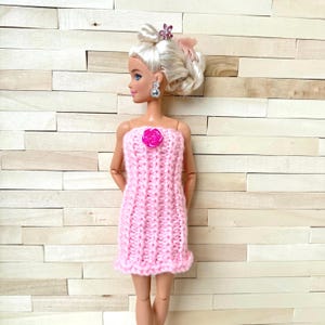 May include: A doll wearing a handmade pink knitted dress with a rose detail. The dress is a tube top style with a ruffled hem. The doll has blonde hair styled in an updo with a pink bow and a jeweled hair accessory. The doll is wearing silver earrings.