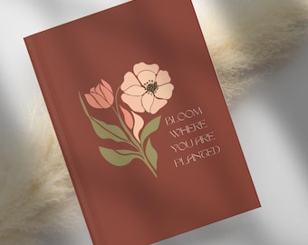 May include: A terracotta-coloured journal with a floral illustration in peach, pink, and green. The journal cover features the text "Bloom Where You Are Planted" in a serif font. The design is minimalist and elegant.