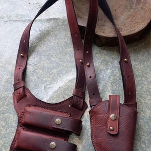 Puede incluir: Una funda de hombro de cuero marrón con dos bolsas para cargadores y una funda para pistola. La funda tiene correas ajustables y remaches metálicos decorativos. El cuero tiene un color marrón rojizo intenso y un acabado texturizado.