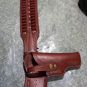 Puede incluir: Un juego de cinturón y funda de cuero burdeos. El cinturón presenta presillas para munición y está adornado con remaches plateados. La funda está diseñada para sujetar una pistola y se asegura con una correa y un botón color latón.
