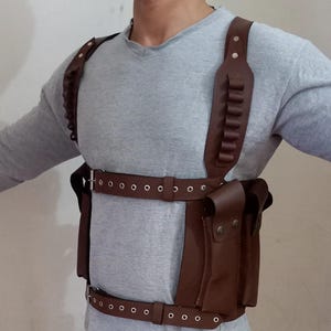 May include: Brown leather chest harness with multiple straps, pouches, and bullet holders. The harness features adjustable straps and a buckle closure. The leather has a rich, dark brown color, and the design appears to be for carrying items.