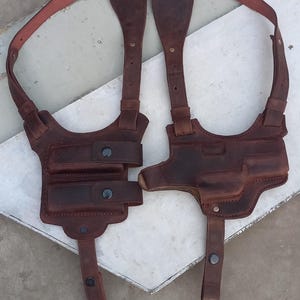 May include: A dark brown leather shoulder holster system featuring two side-by-side pouches on one side and a moulded holster on the other. The harness includes adjustable straps with metal rivets and snap button closures for secure storage.