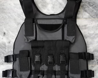 May include: Gray tactical vest with multiple black pouches and adjustable straps. The vest features several compartments secured with buckles and hook-and-loop closures. The vest is designed for carrying equipment.