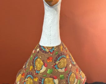 May include: A white goose statue wearing a brown fabric outfit featuring a repeating pattern of turkeys and colorful autumn leaves. The outfit includes a matching bow attached to the goose's head, with a red ribbon and lace trim along the bottom hem.