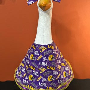May include: A white decorative goose figurine wearing a purple and yellow fabric outfit featuring the LSU Tigers logo and football helmet graphics. The goose also wears a matching purple and yellow bow on its head, with a yellow lace trim.
