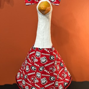 May include: A white goose figurine wearing a red fabric outfit and a matching bow, both patterned with the Georgia Bulldogs logo, the word UGA, and the letter G. The goose has a yellow-orange beak and is dressed in a flared, skirt-like garment.