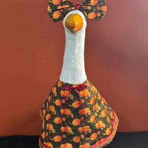 May include: A decorative goose figurine with a white neck and head, a yellow beak, and a bow tie and dress featuring a pumpkin pattern. The dress has a red ribbon and lace trim, and the base is green.