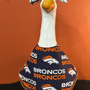May include: A white goose statue wearing a dark blue fabric outfit featuring the Denver Broncos logo and the word BRONCOS in orange and white. The goose also wears a matching fabric bow tie on its head, and the outfit has an orange lace trim.