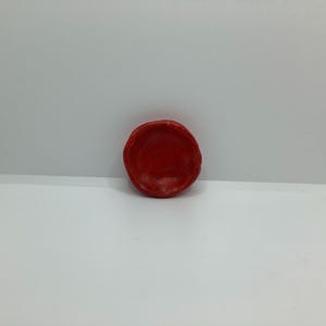May include: A small, round, red ceramic bowl with a textured surface. The bowl is centered against a white background. The bowl's shape is slightly irregular, with a rough, handmade appearance.