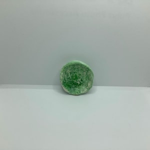 May include: A round, green soap bar with a textured surface. The soap has a white base and a vibrant green top layer, creating a marbled effect. The soap is a solid, circular shape.