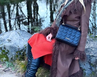 Può includere: Un cappotto lungo marrone con finiture in ecopelliccia, una borsa nera con un motivo a cuore, una gonna rossa e stivali neri al ginocchio. L'outfit è completato da una sciarpa e un cappello, creando un look alla moda.